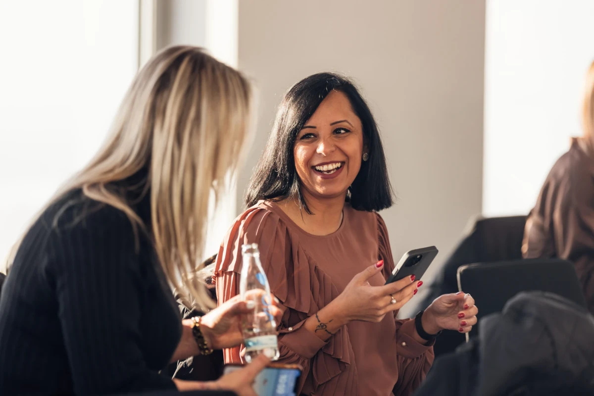In dem Bild sieht man mittig eine Frau, mittellange schwarze Haare mit einem Handy in der Hand. Sie l&auml;chelt die Person links von ihr an. Die linke Person ist mit dem R&uuml;cken zu der Kamera gedreht. SIe hat blonde lange Haare, h&auml;llt ebenfalls ein Handy in der Hand und eine Trinkflasche. | In the center of the picture you can see a woman with medium-length black hair holding a cell phone. She is smiling at the person to her left. The person on the left has their back to the camera. She has long blonde hair and is also holding a cell phone and a water bottle. | SPIRIT/21