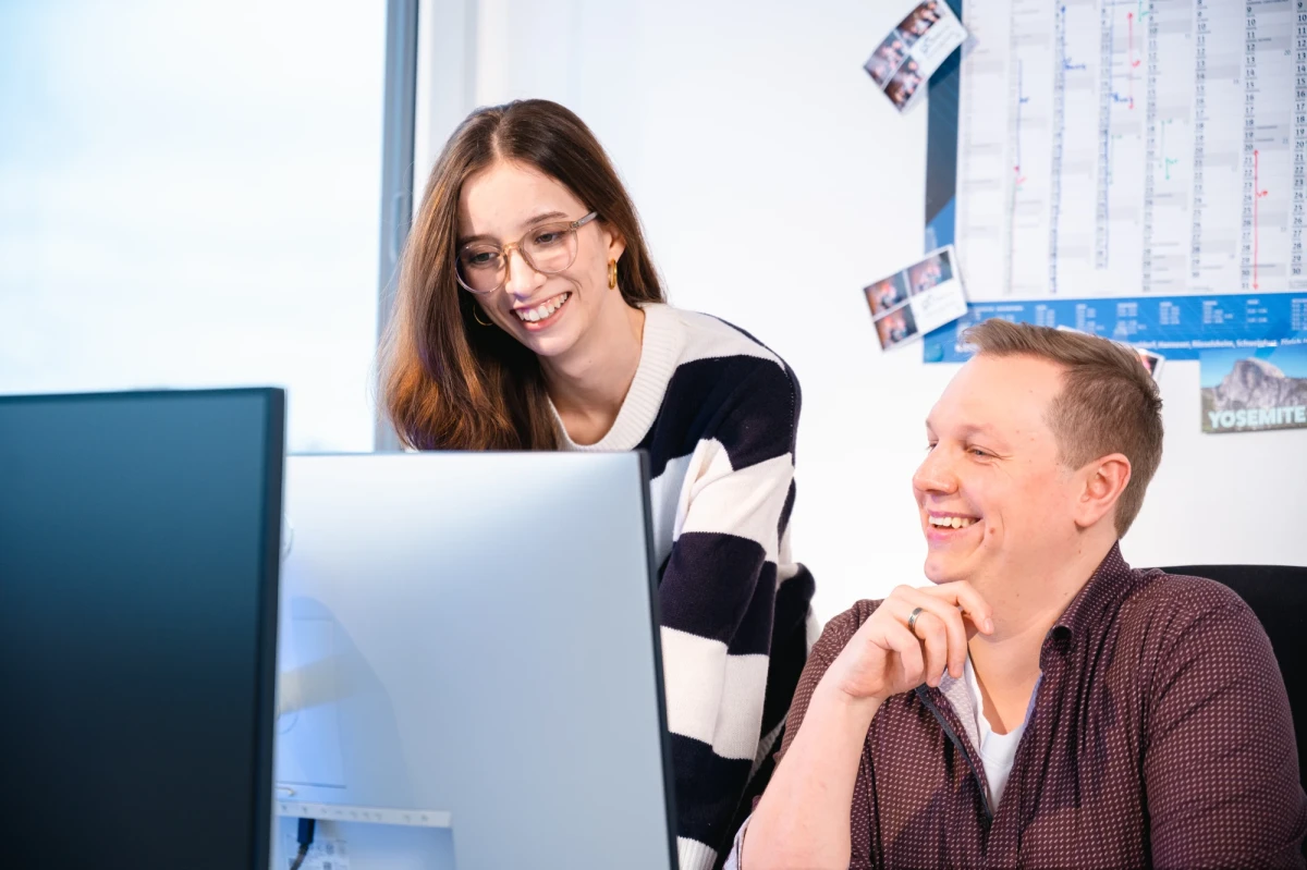 In dem Bild sind 2 Personen zu sehen. Eine Person sitzt vor dem Laptop, die andere Person daneben steht. Sie schauen beide in den Bildschirm der vor Ihnen steht. Die sitzende Person, rechts, ist ein junger Mann. Er hat kurze blonde Haare und l&auml;chelt. Links von ihm steht eine junge Frau. Sie hat mittellange braune Haare, eine Brille und sie l&auml;chelt. | The picture shows 2 people. One person is sitting in front of the laptop, the other person is standing next to it. They are both looking at the screen in front of them. The person sitting on the right is a young man. He has short blond hair and is smiling. To his left is a young woman. She has medium-length brown hair, glasses and is smiling. | SPIRIT/21