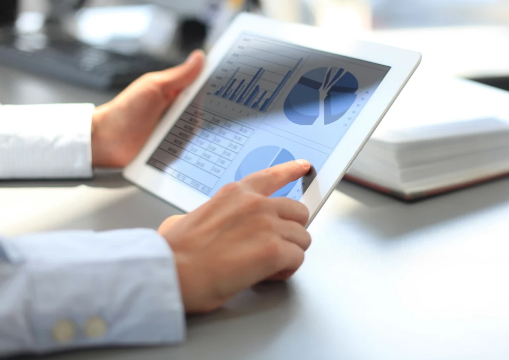 Zwei H&auml;nde, welche in ein IPad halten, auf dem verschiedene Diagramme zu sehen sind. Im Hintergrund sieht man verschiedene B&uuml;rogegenst&auml;nde.